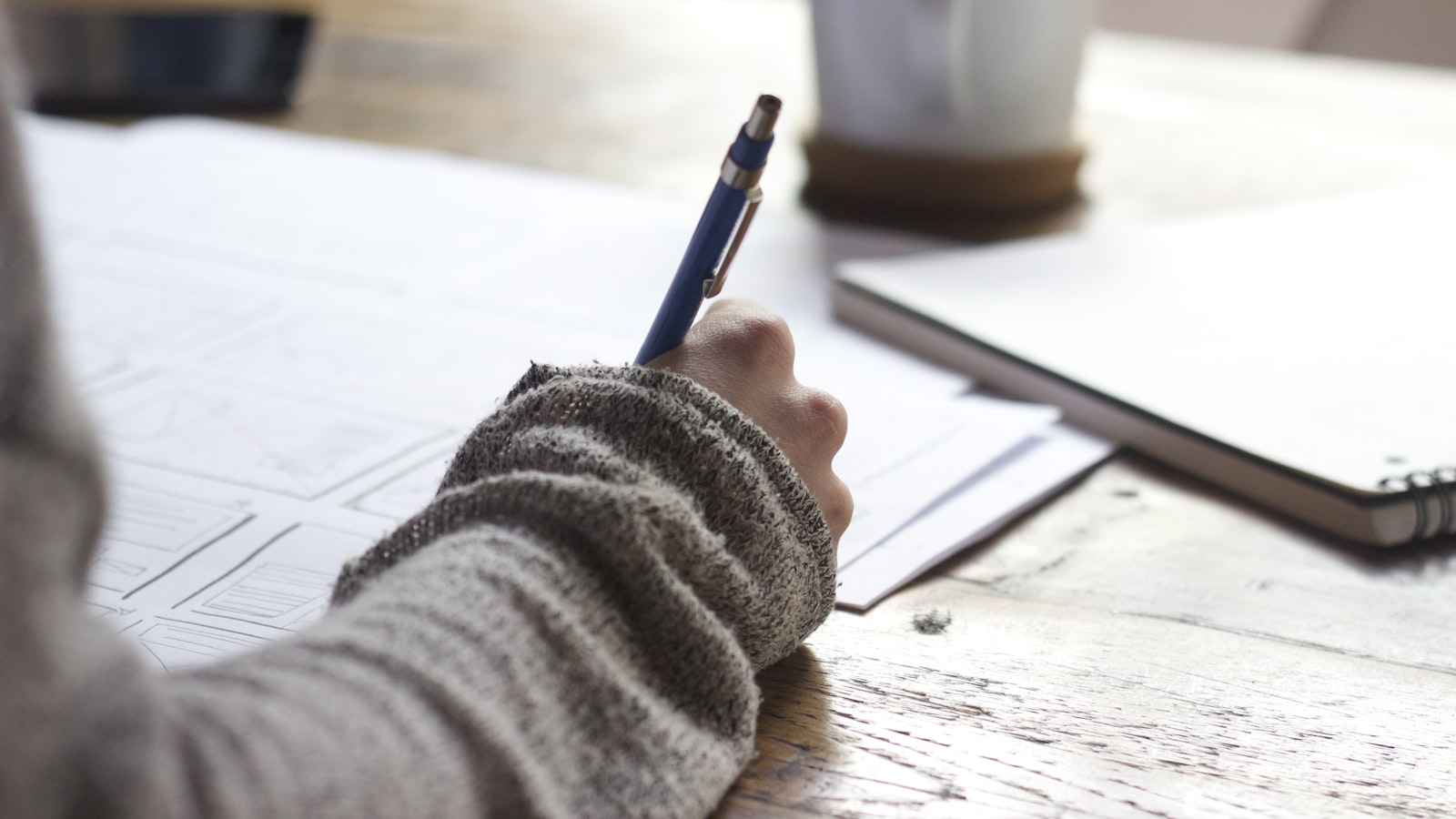 A pair of hands writing notes in an open notebook beside a cup of coffee.